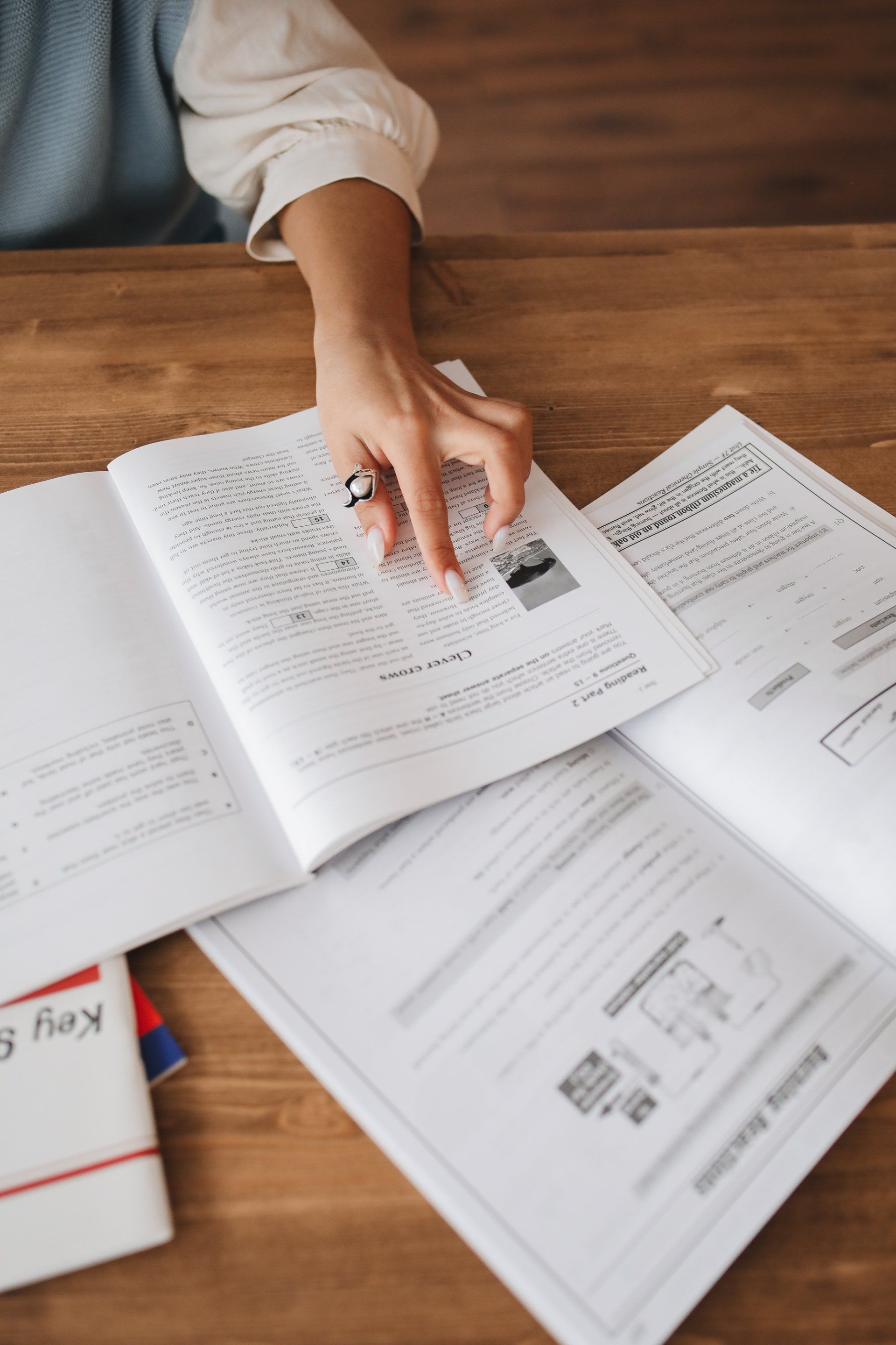 Person's hand pointing at open book on a wooden table, surrounded by other open books and a key guide.