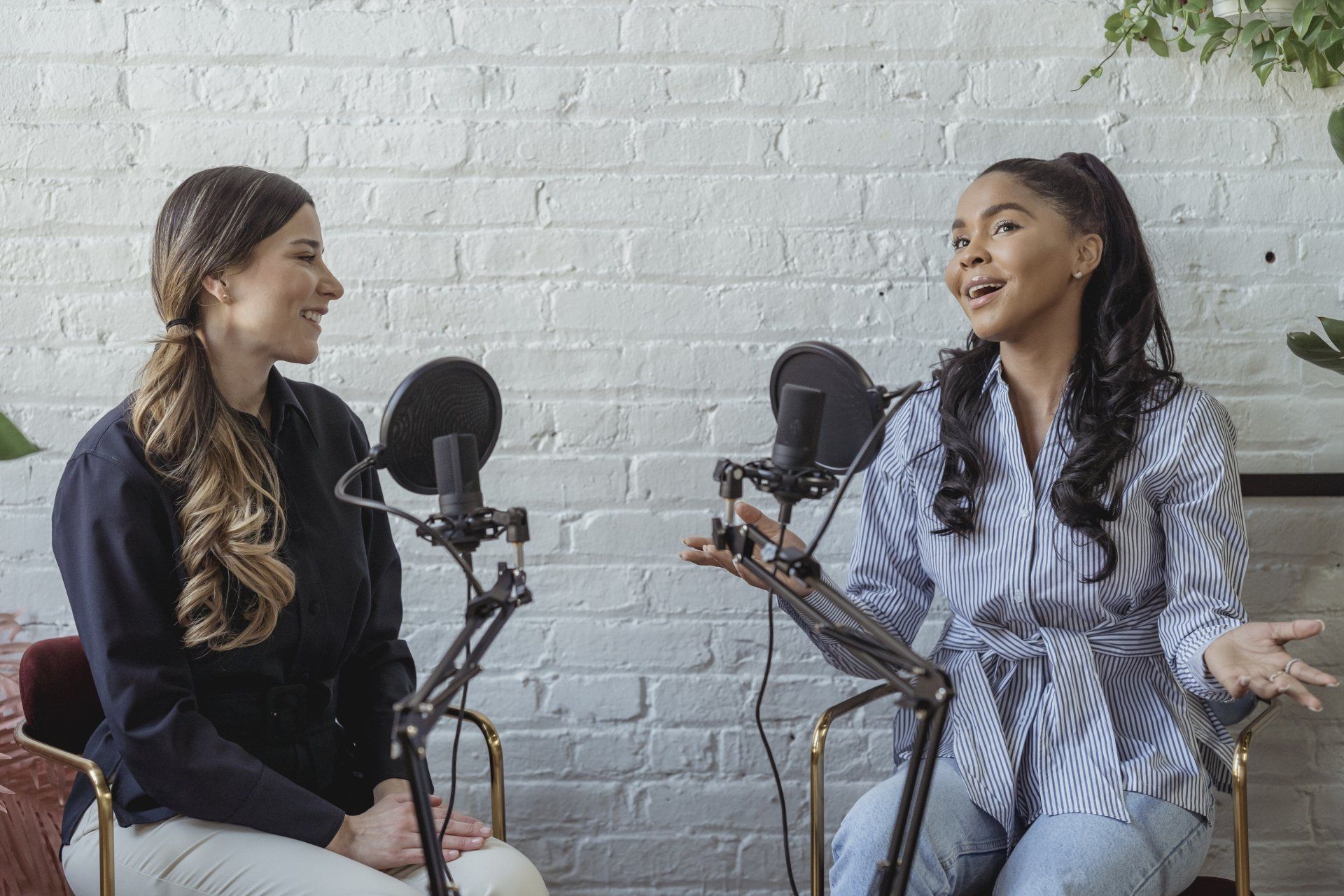 Two women sit at microphones, recording a podcast in front of a white brick wall.