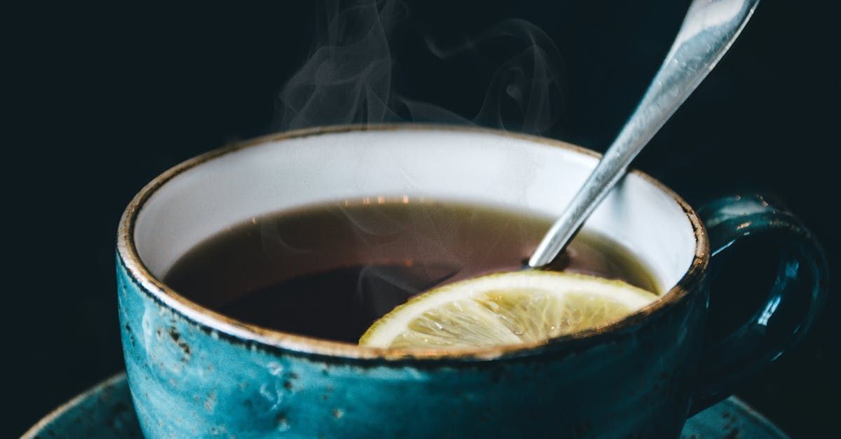 Blue ceramic mug with hot tea, a lemon slice, and a spoon, steaming against a dark background.