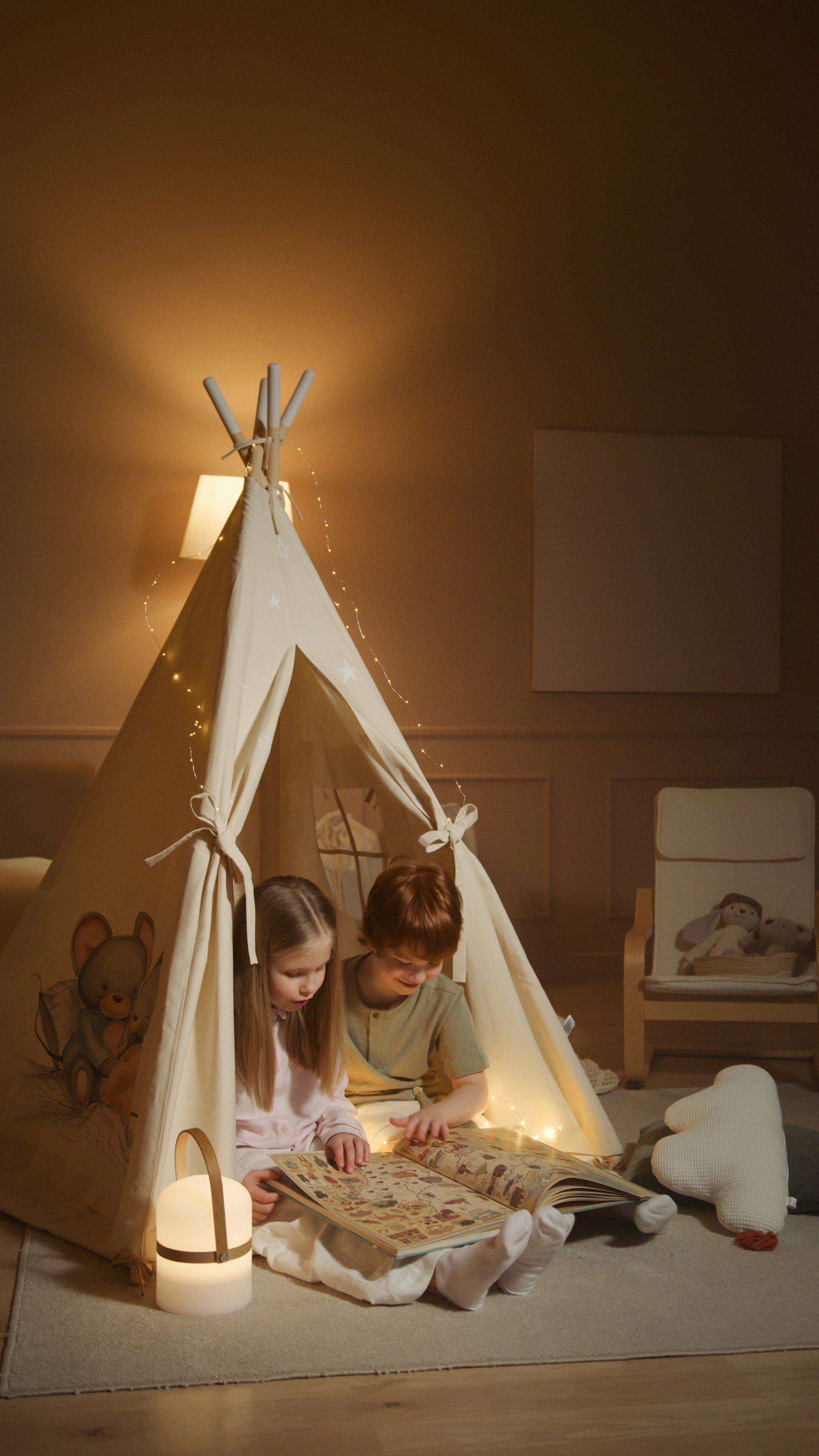 Two children reading inside a lit-up teepee tent. Beige interior, fairy lights, and a lamp.