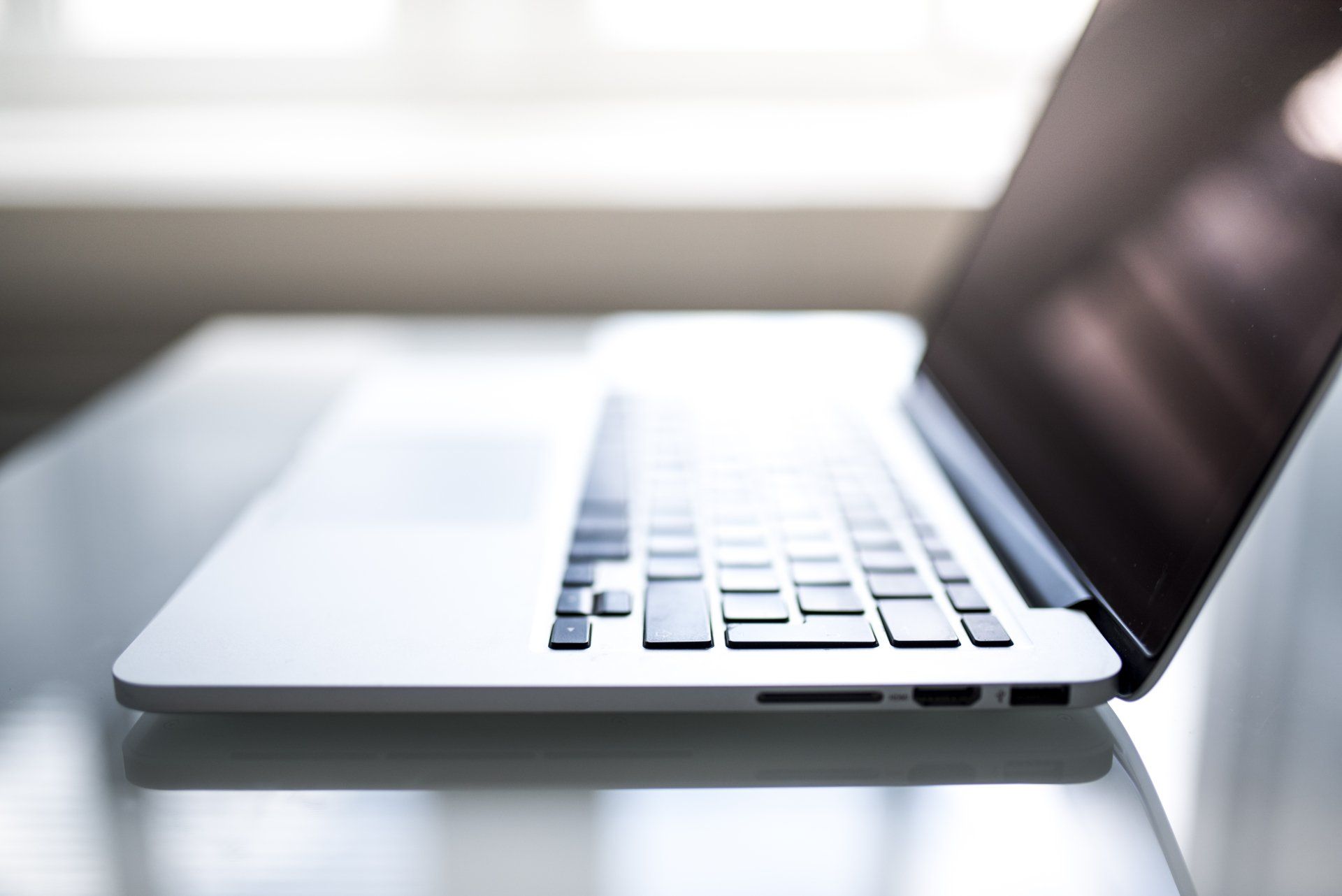 Silver laptop on a glass table, partially open with the screen facing away, near a window.