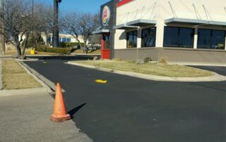 An orange traffic cone is sitting on the side of the road in front of a burger king restaurant.