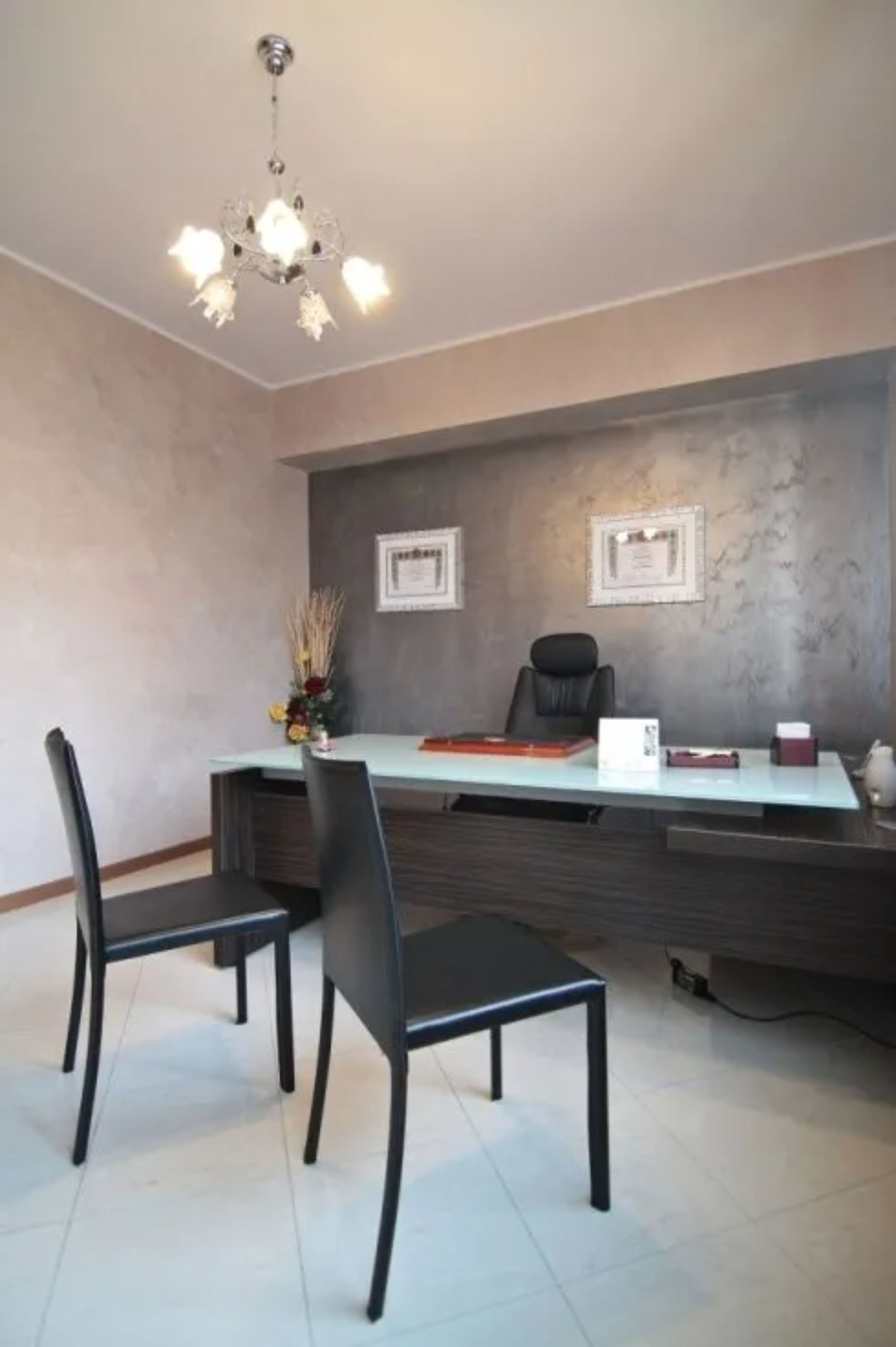 Ufficio moderno con una grande scrivania, una sedia in pelle nera, due sedie nere e un lampadario decorativo.