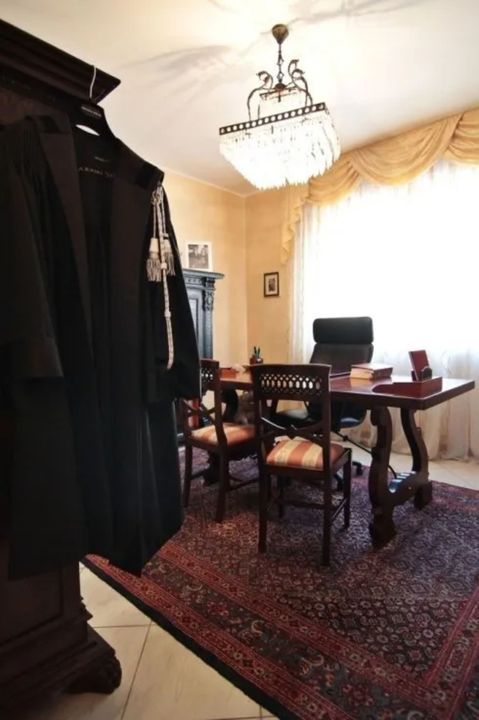 Ufficio con una grande scrivania, una sedia in pelle nera, due sedie nere per i visitatori e un lampadario decorativo.