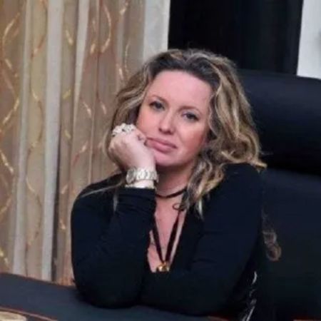 Donna con i capelli biondi, vestita di nero, con il mento appoggiato sulla mano, che guarda la telecamera in una stanza.