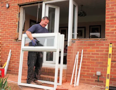 Man installing a white window in a brick wall exterior. He is wearing blue gloves.