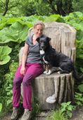 Frau und Hund sitzen auf einem geschnitzten Holzstumpf in einem Wald. Die Frau trägt lila Hosen und lächelt, der Hund ist schwarz und weiß.