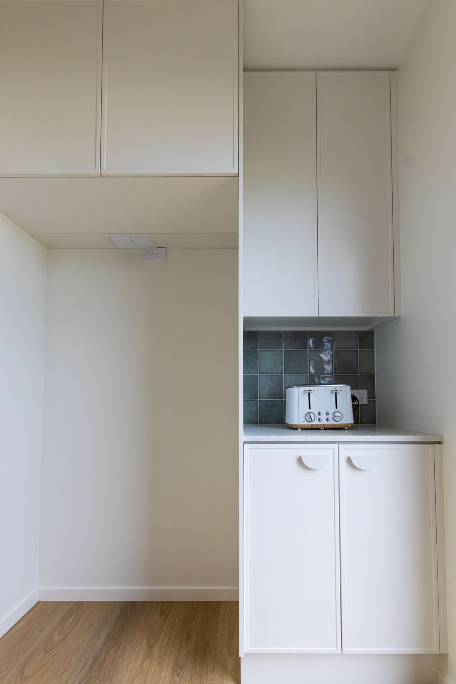 A small, white kitchenette with upper cabinets, a countertop, and a toaster on a tiled backsplash.