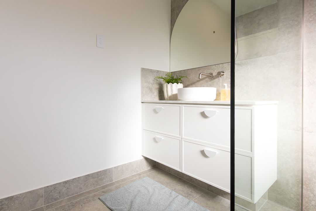 White bathroom vanity with a round mirror, small plant, and gray tile flooring.