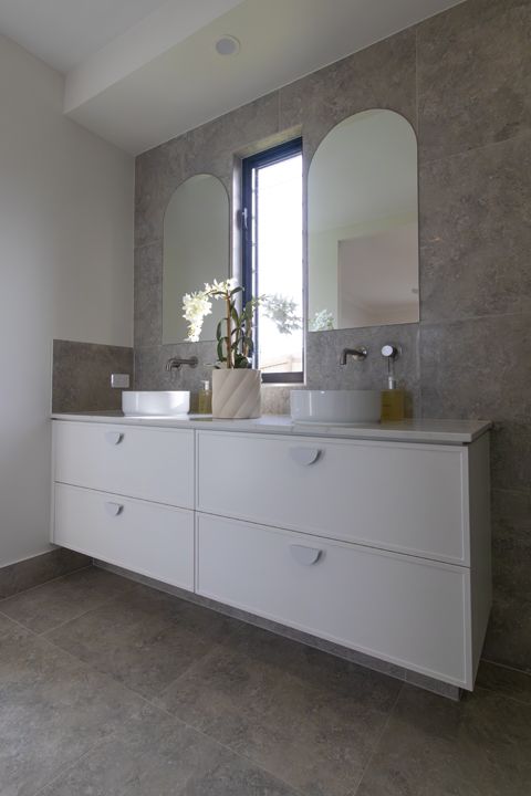 Bathroom with white floating vanity, two sinks, arched mirrors, and gray tile.