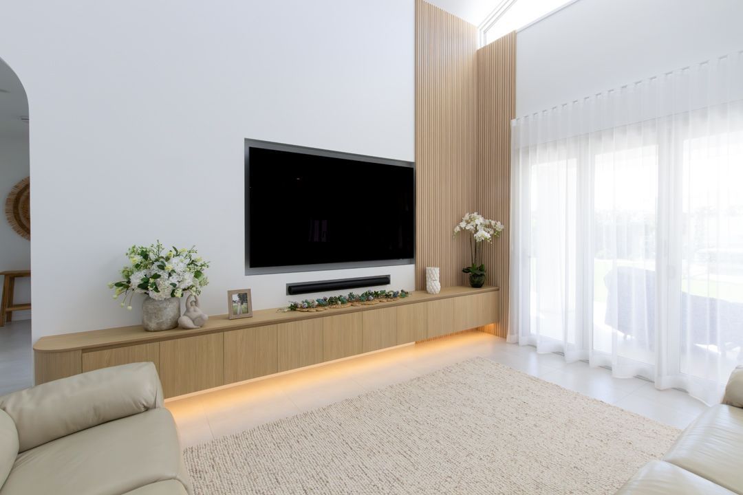 Living room with mounted TV, wooden cabinet, large rug, and beige furniture. Bright, natural light through sheer curtains.