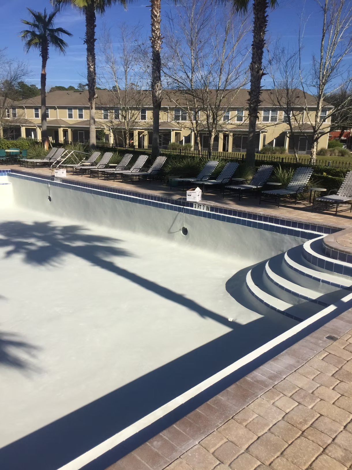 Empty swimming pool with blue trim, lounge chairs, and a sunny apartment courtyard in the background