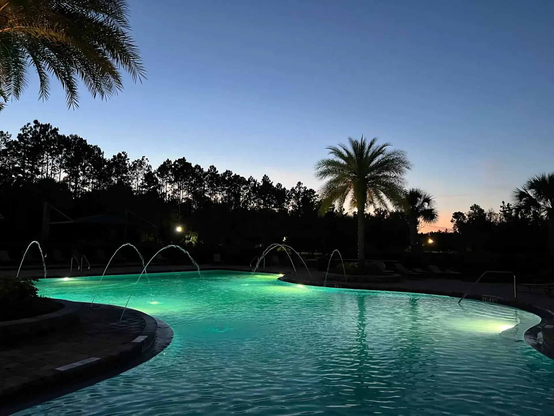 Curved backyard pool glowing turquoise at dusk with palm trees silhouetted against a blue-orange sky