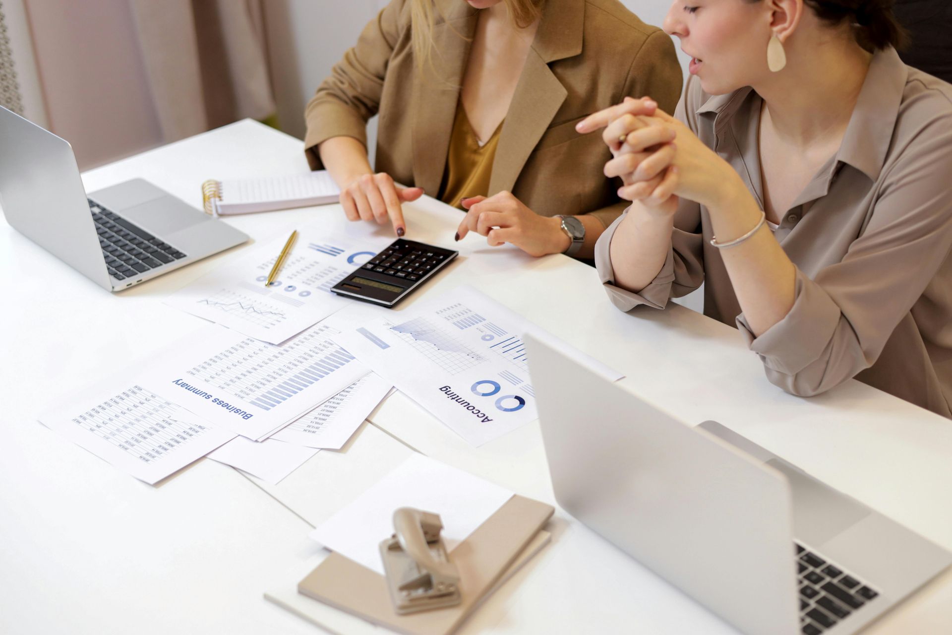 Two women at a desk, reviewing papers, working on laptops, and using a calculator in an office.