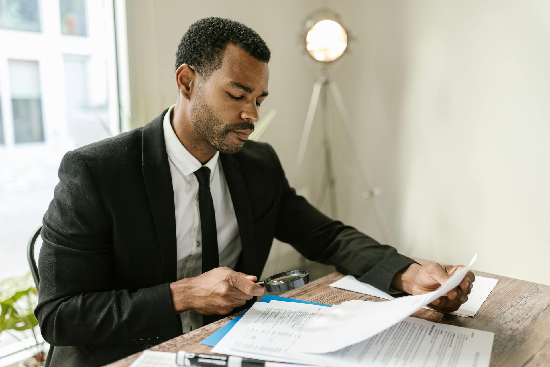 Man in suit reviewing documents with a magnifying glass at a desk.