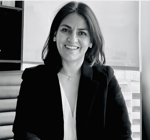 Una mujer con blazer negro sonríe; lleva camisa blanca y joyas. Interior, cerca de una ventana con persianas.