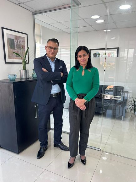 Un hombre y una mujer en una oficina. El hombre lleva traje. La mujer lleva camisa y pantalones verdes.