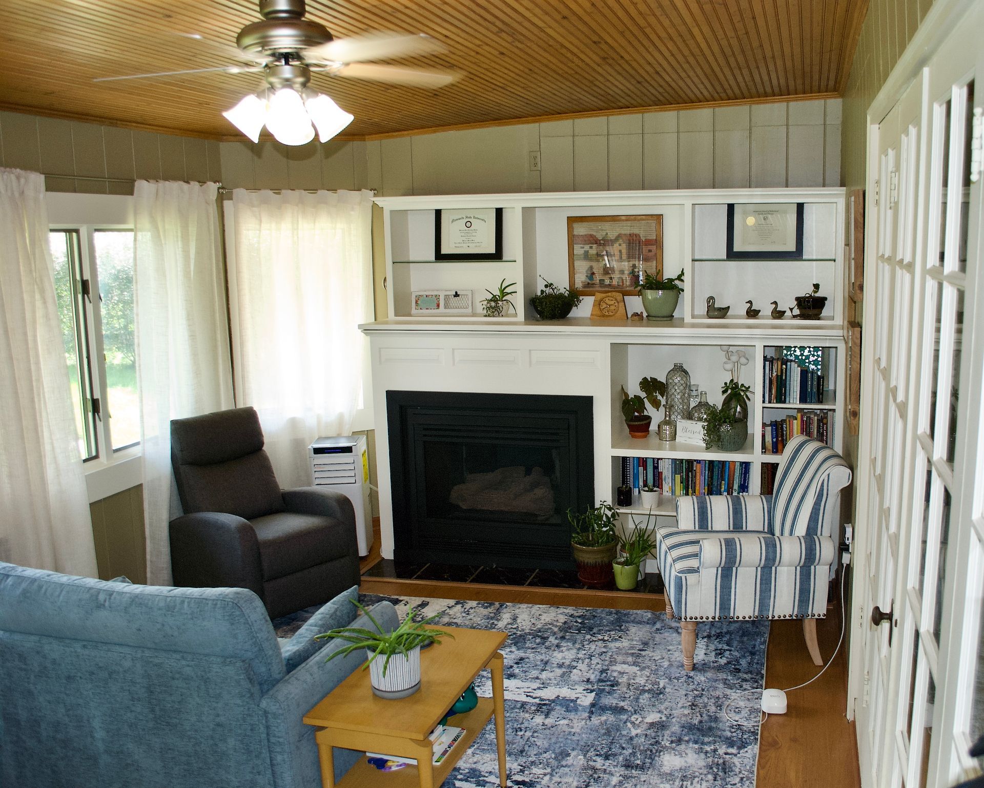 Cozy living room with fireplace, built-in shelves, blue furniture, white curtains, and wood ceiling.
