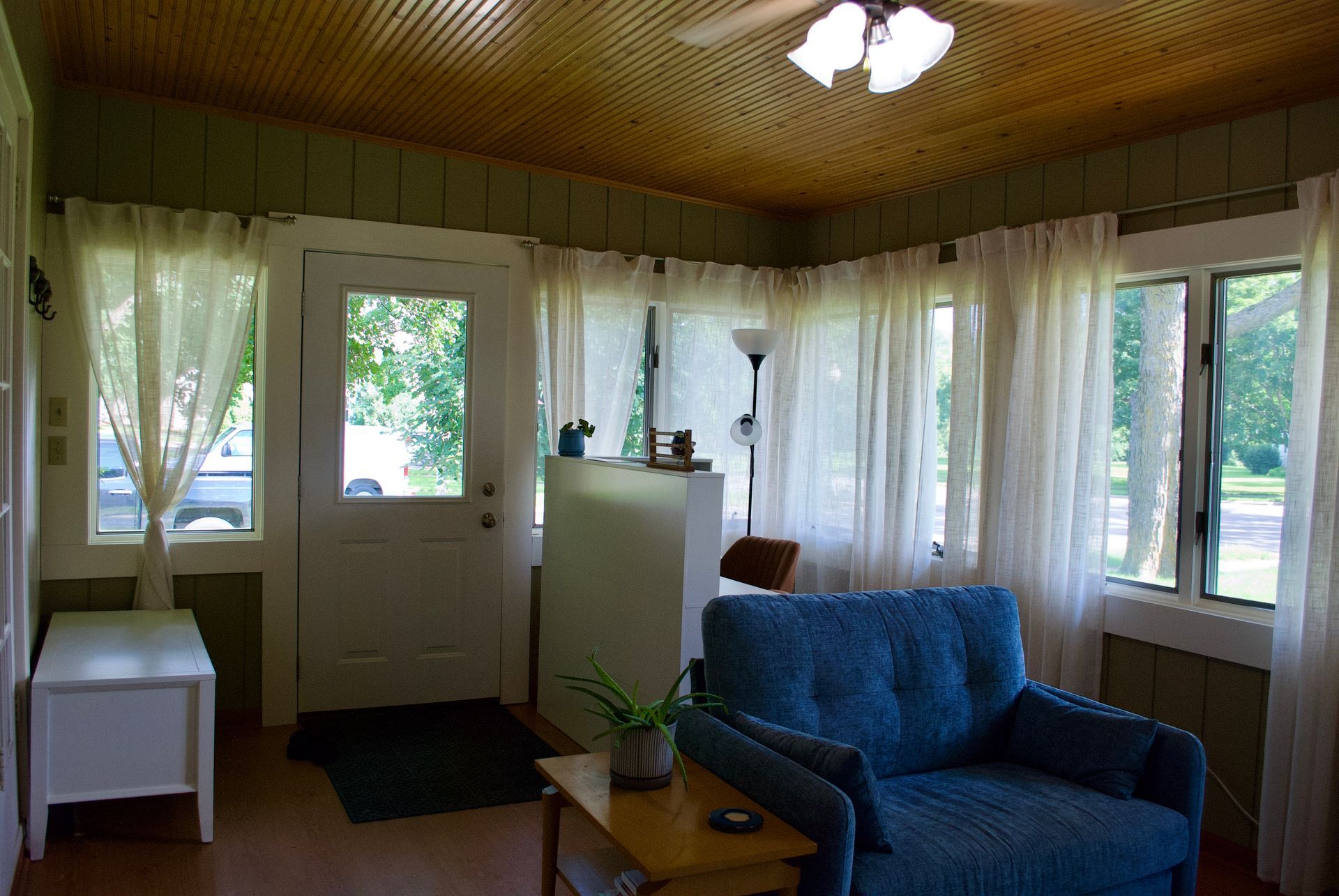 Sunroom with white curtains, blue armchair, wood ceiling, and door to outside.