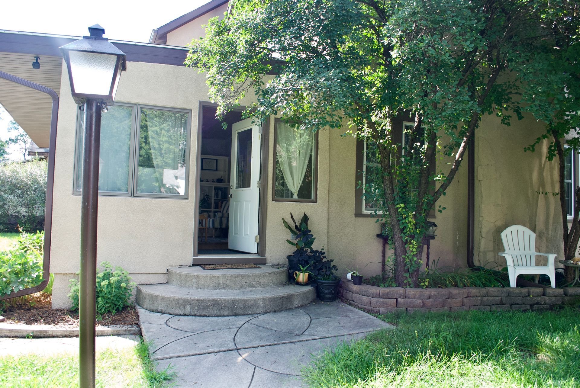 Exterior of a small house with a curved concrete walkway, front door open, small porch and lamp.