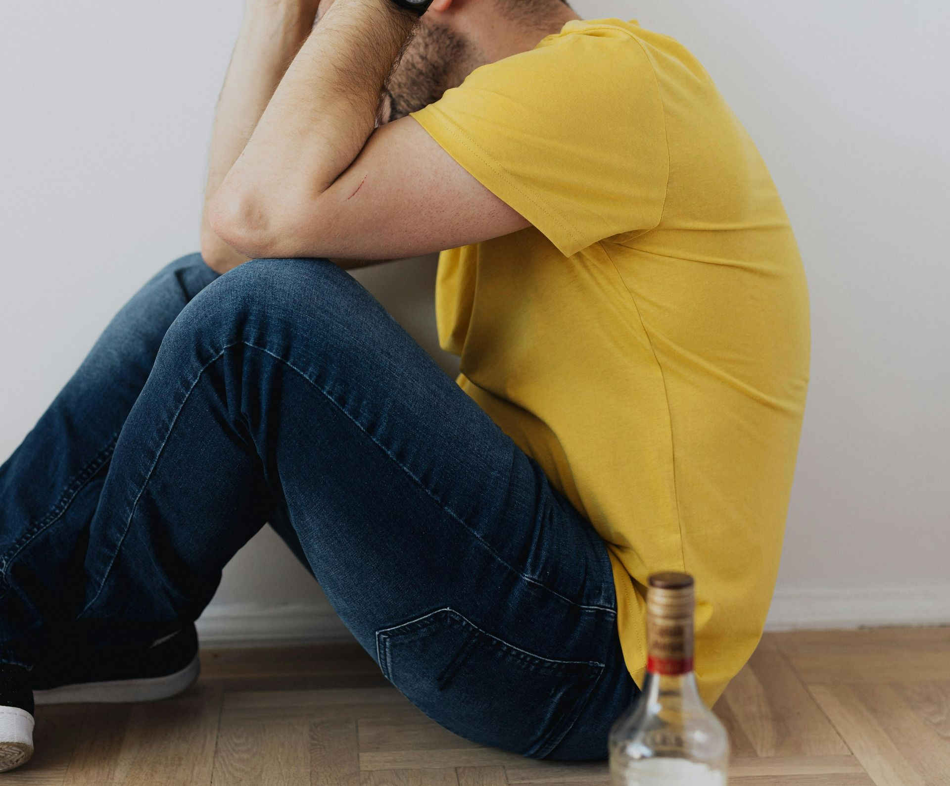 Man in yellow shirt and jeans, sitting against a wall, head in hands, bottle in the foreground.