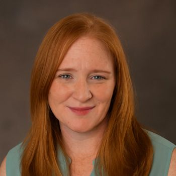 Woman with long red hair smiles, wearing a turquoise top, against a blurred background.