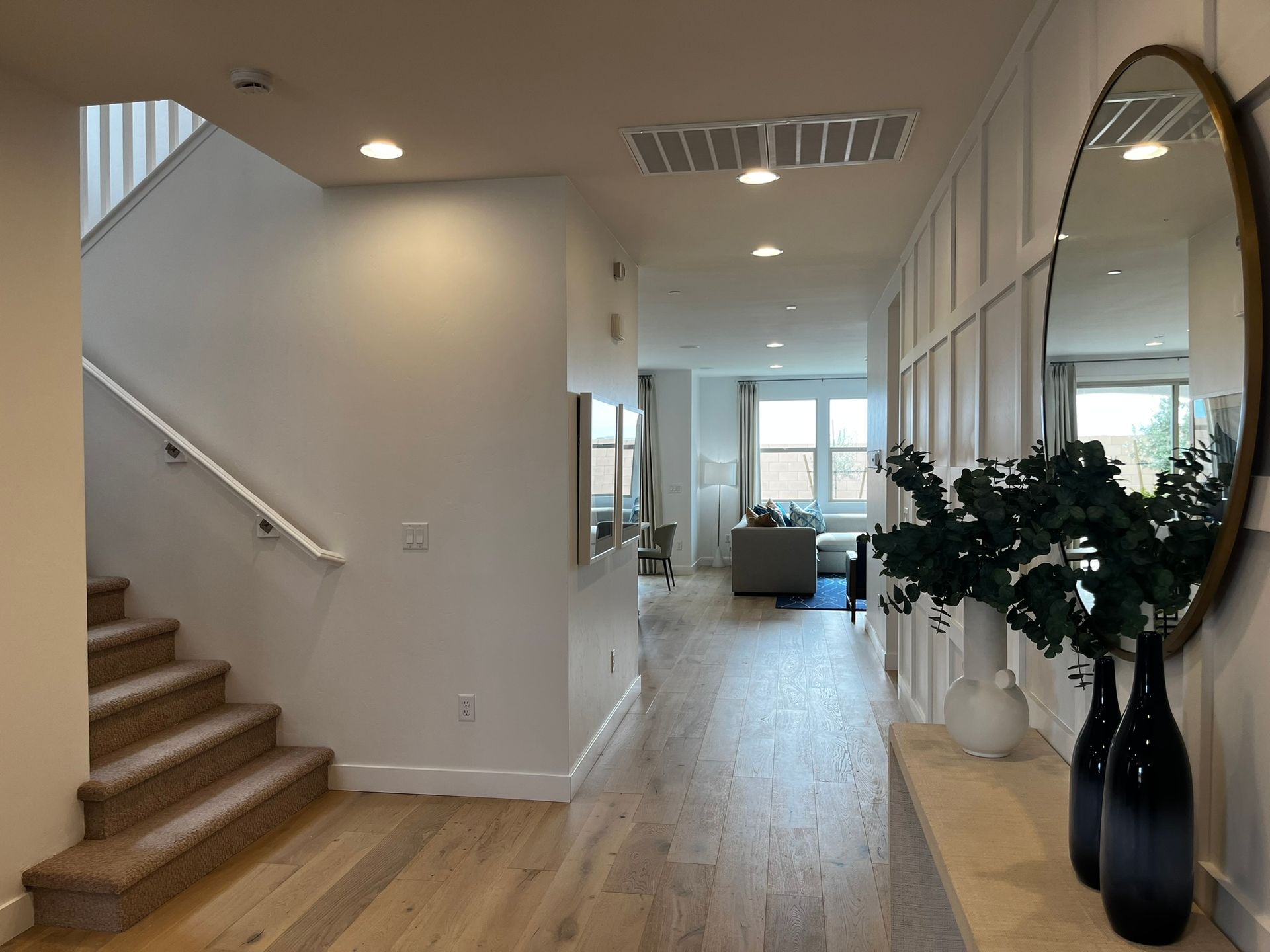 A modern home interior featuring a hallway with wood flooring, a staircase, and a decorative mirror with plants.