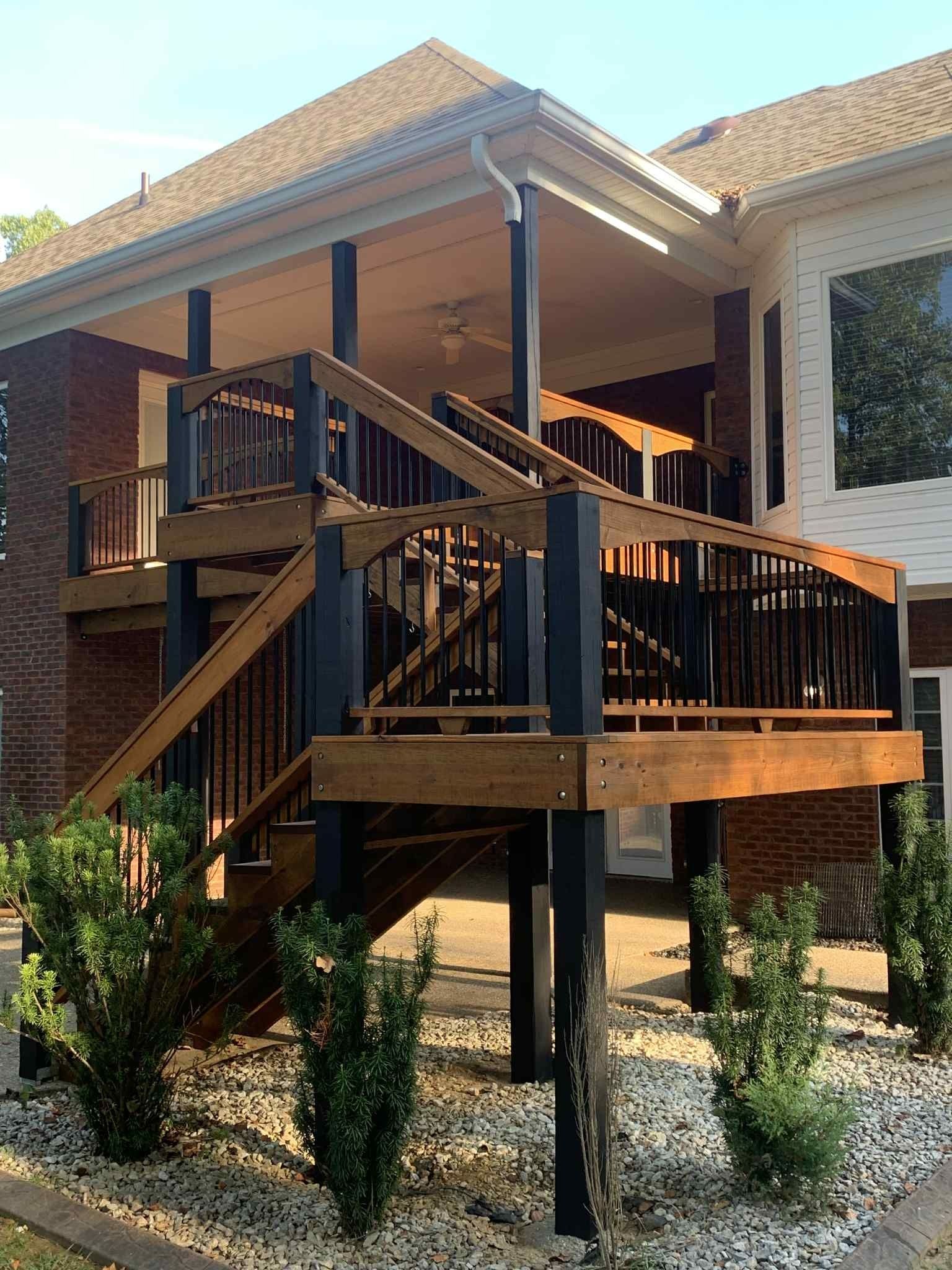 Two-level wooden deck with black railings and columns attached to a brick house, small bushes in front.