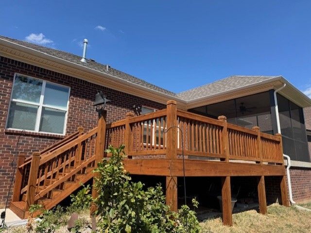 Wooden deck stained brown attached to a brick house under a blue sky.