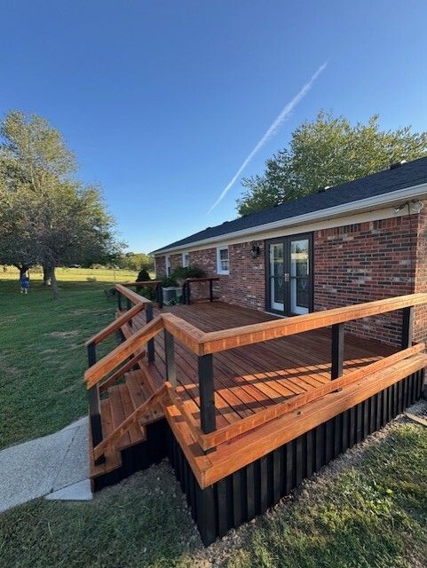 Wooden deck attached to a brick house. Steps lead down to the grassy yard. Blue sky.