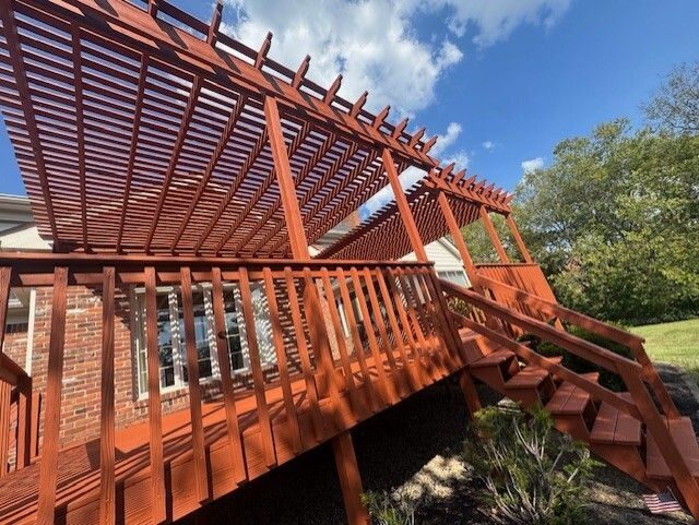 Red wooden deck and pergola against a blue sky, attached to a house with a green lawn.