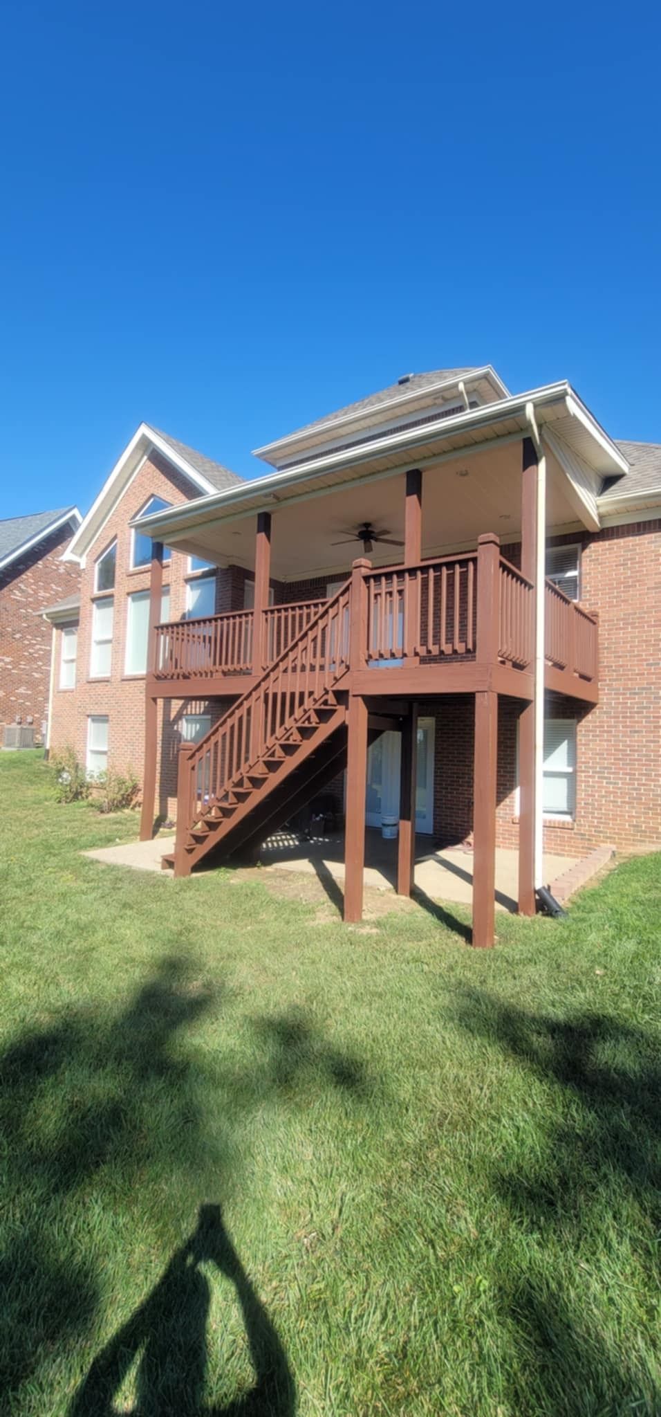 Two-story brown deck attached to a brick house on a green lawn, under a bright blue sky.