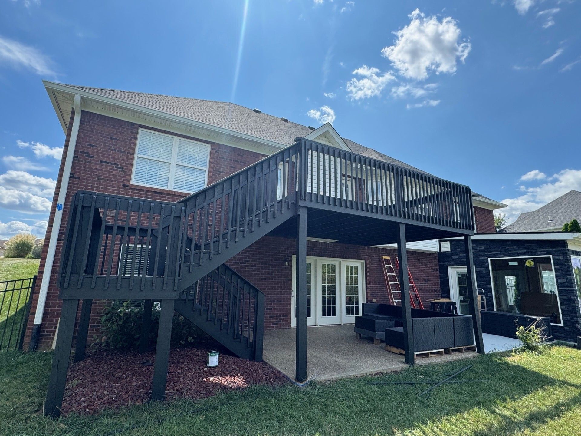 Dark wood deck attached to a brick house with white trim. Blue sky and green grass.