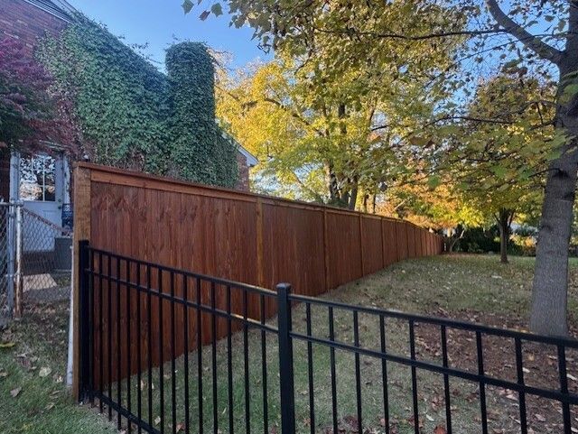 Wooden privacy fence with black metal fence in front; beside a brick building with vines.