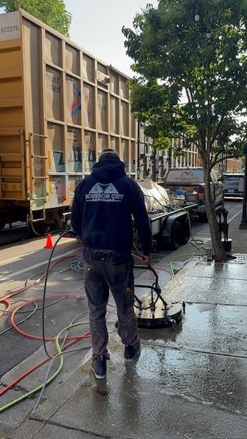 Person power washing a sidewalk in front of a train car, street setting.