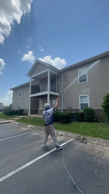 Person power washing the side of a beige building with a high-pressure hose on a sunny day.
