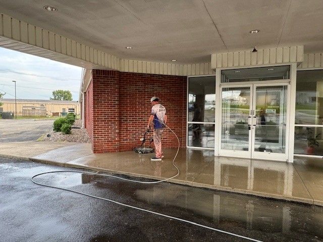 A person power washes a brick wall and concrete sidewalk outside a building with glass doors.