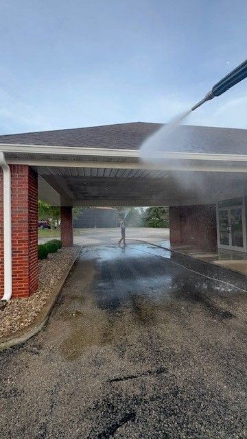 A person power washing a building's roof overhang; brick and concrete exterior.