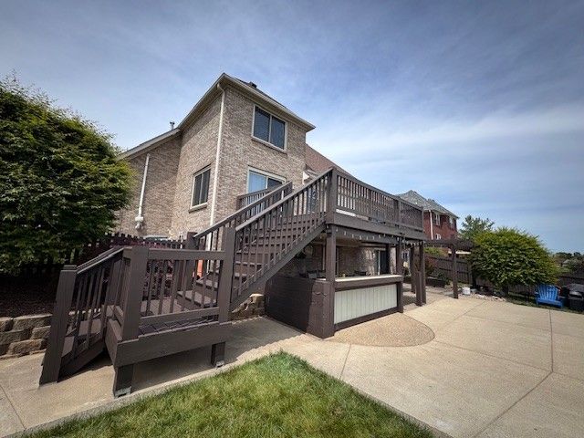 Brown wooden deck with stairs attached to a brick house. Green lawn and a sunny, cloudy sky.