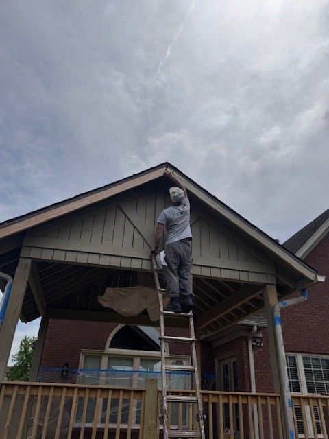 Person on ladder painting the trim of a covered porch on a brick house. Blue tape is visible.