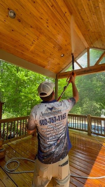 Person power washing wooden porch ceiling and deck. Green trees in background.