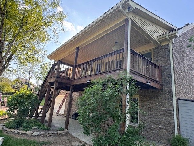 Wooden deck with stairs attached to a two-story brick house, surrounded by green shrubs and trees.