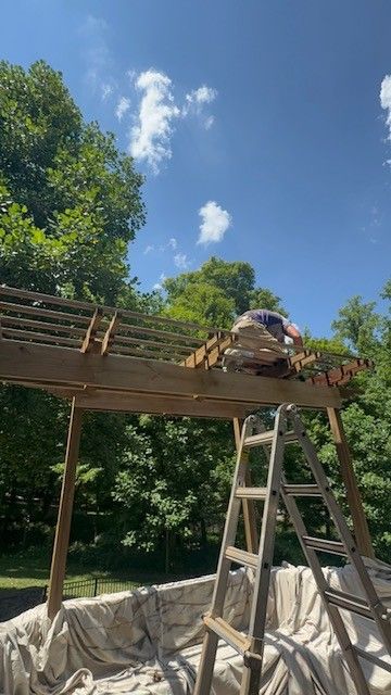 Person working on wooden pergola roof with a ladder, blue sky, and trees.