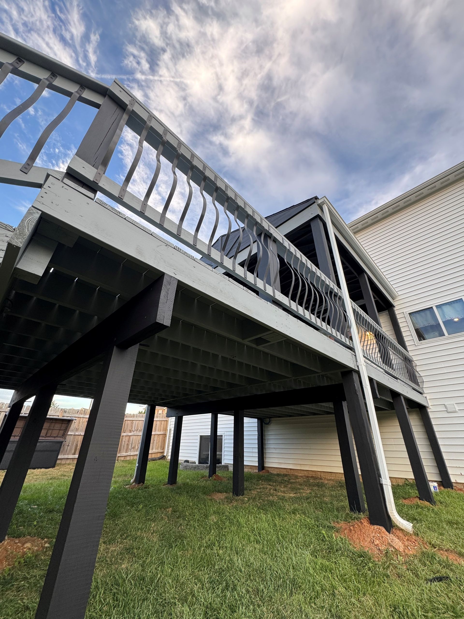 Low-angle view of a gray deck with black supports on green grass, next to a white house under a cloudy sky.