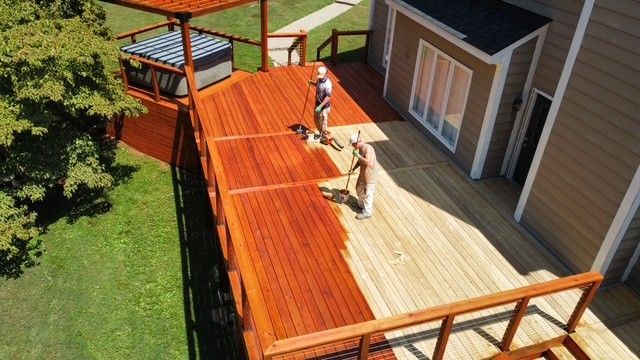 Two people staining a wooden deck red, next to a beige house, with a hot tub and pergola.