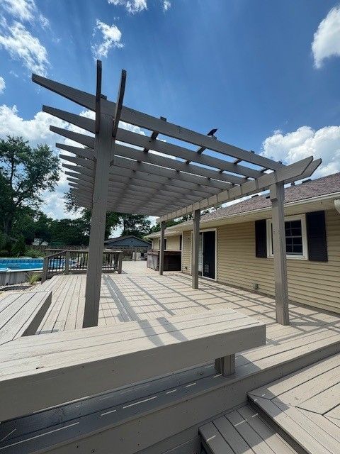Wooden pergola on a deck with a swimming pool and house in the background on a sunny day.