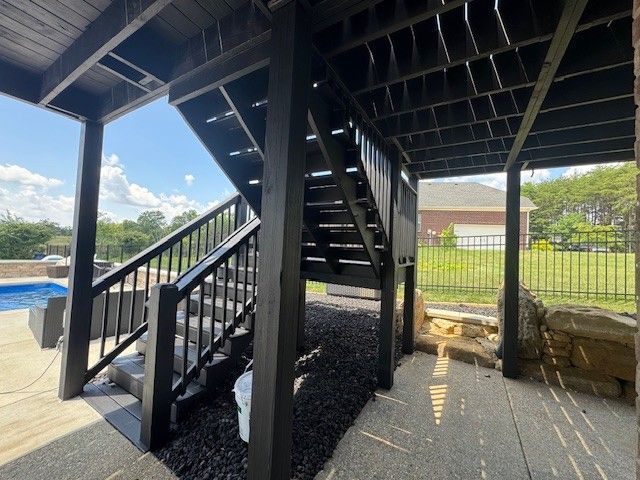 Black-painted wooden deck with stairs leading down, overlooking a pool and yard with a fence.