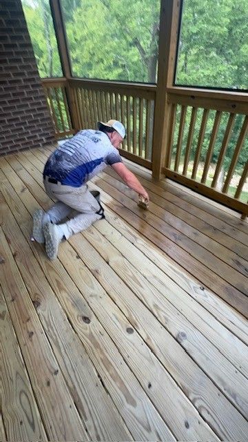 Person kneeling on a deck, applying stain with a brush. Wooden railing, green trees visible.