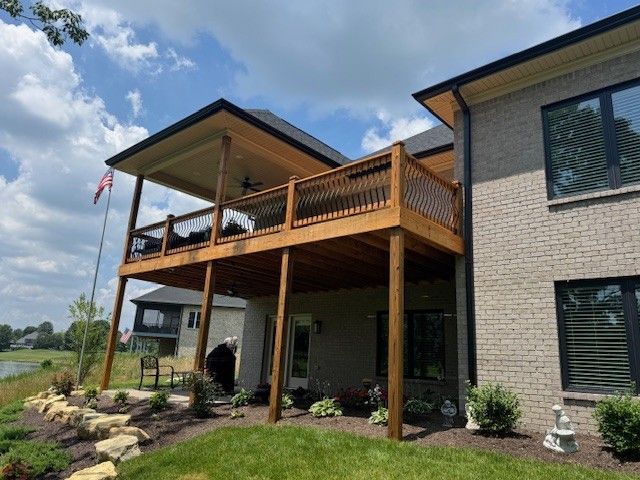 Two-story wooden deck attached to a brick house. Includes an outdoor ceiling, railing, and support beams. Overlooks a yard.