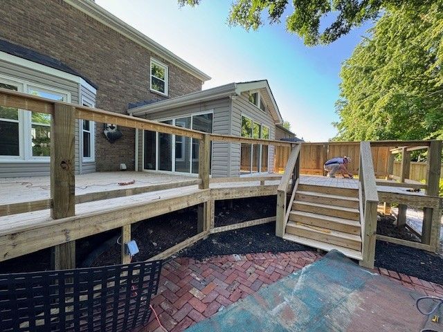 Wooden deck attached to a two-story brick house with stairs, a railing, and a view of a sunroom.